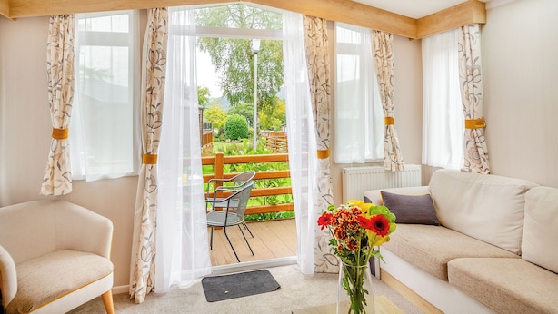 Een kamer met een houten plafond, een bank, een stoel en uitzicht op een tuin door grote ramen.