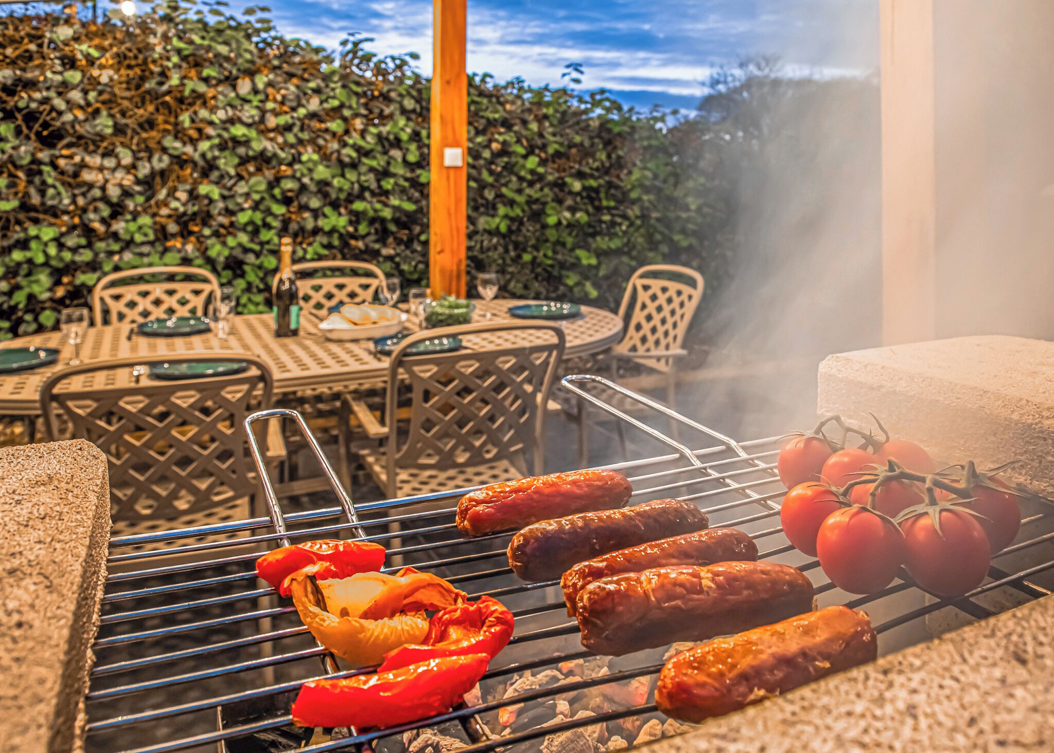 Grill med pølser og tomater, udendørs møbler og en hæk i baggrunden.