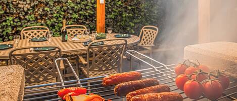 Grill med pølser og tomater, udendørs møbler og en hæk i baggrunden.