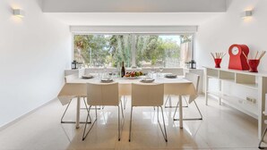 A modern dining area with a large table set for four, white chairs, and a view of greenery through large windows. - Palmera Sky Terrace Suite (Costa del Silencio)