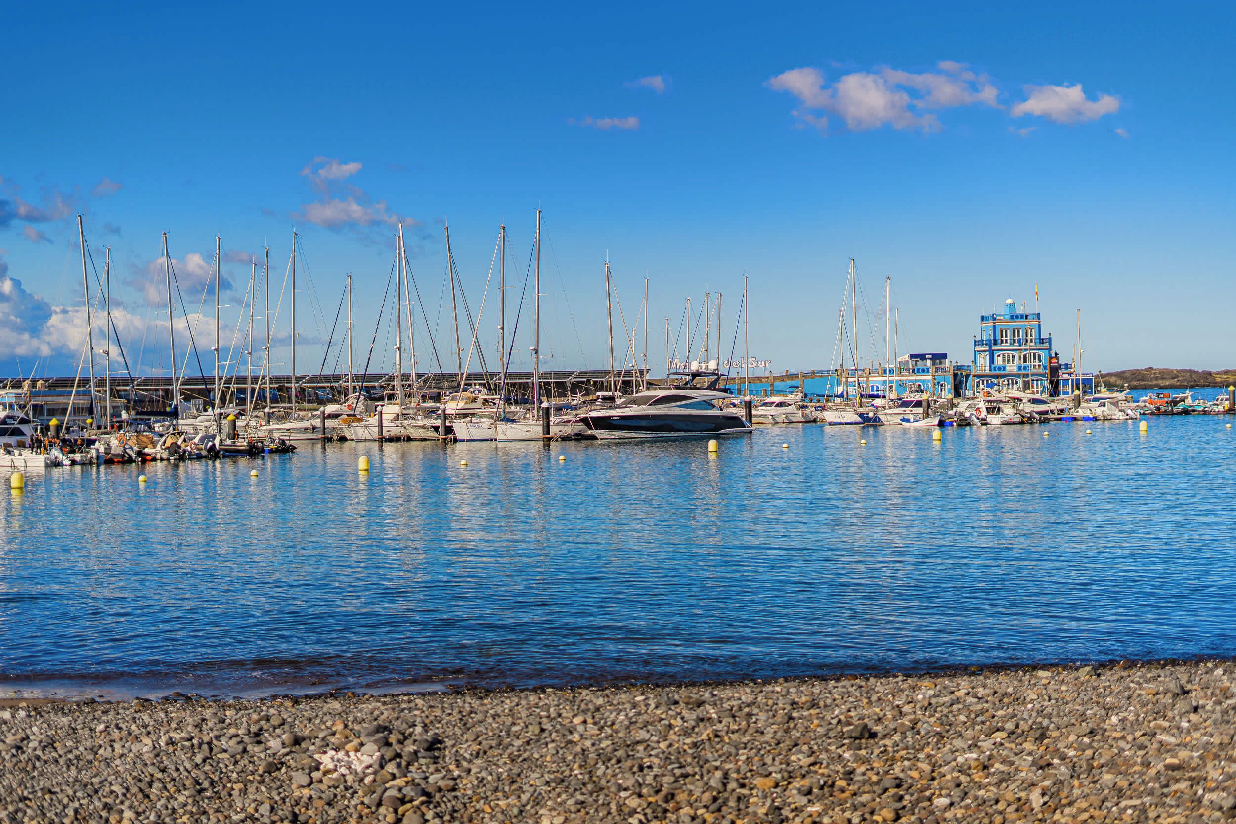 A marina with boats docked, a building with a blue facade, and a rocky shoreline.