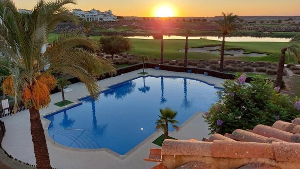 Piscina com vista para o pôr do sol, rodeada por palmeiras e um campo de golfe ao fundo.