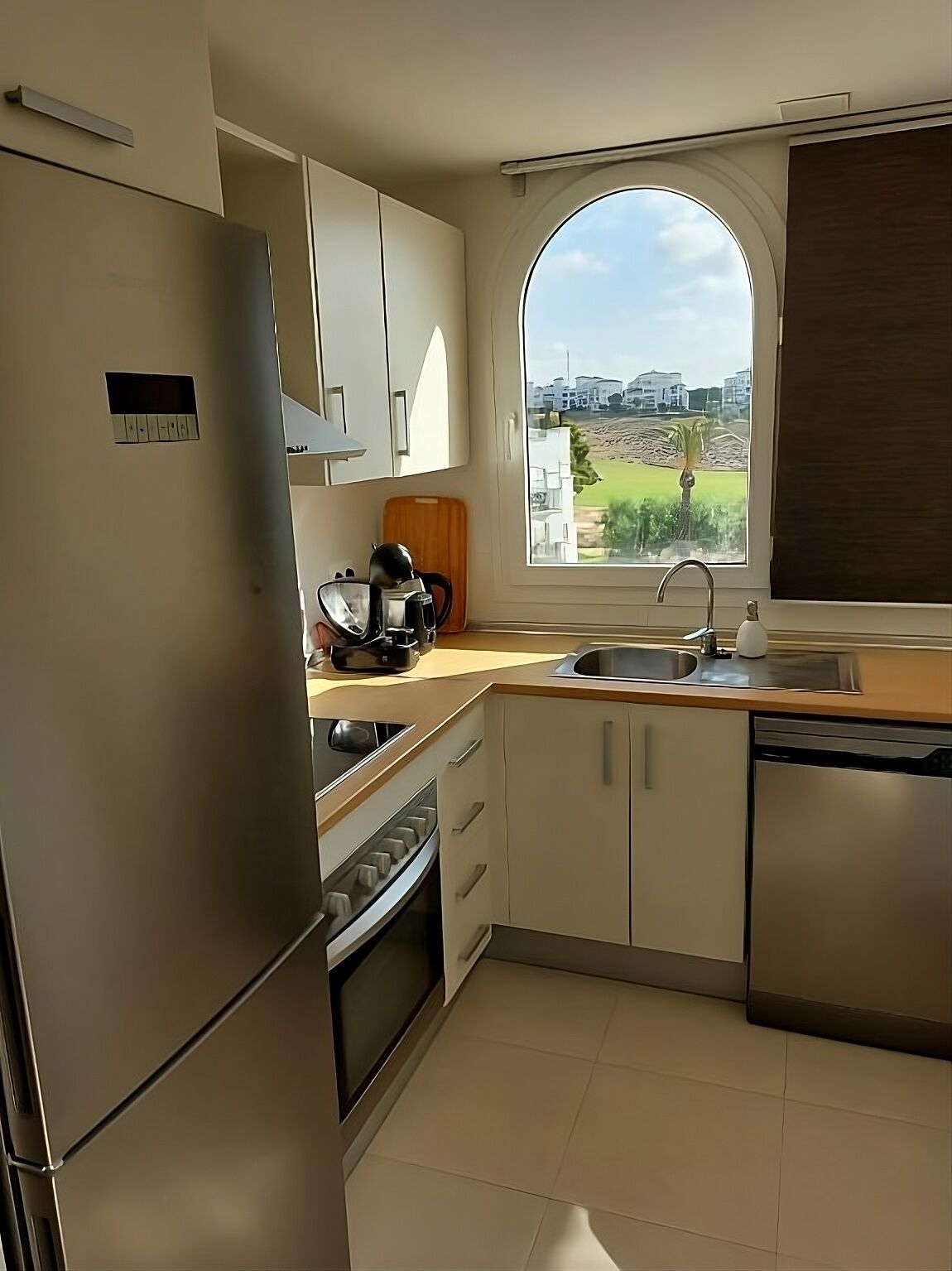 Cozinha moderna com geladeira de aço inoxidável, forno, pia e janela com vista para uma área residencial.