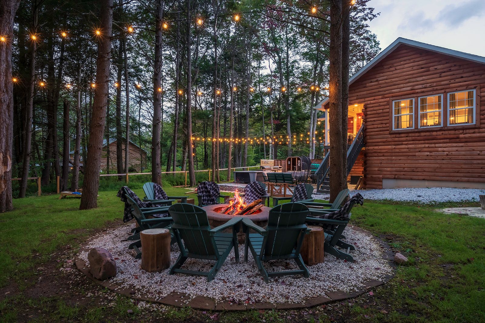 Eine Blockhütte mit einem Feuerkorb, umgeben von Stühlen, Lichterketten und einem Wald im Hintergrund.