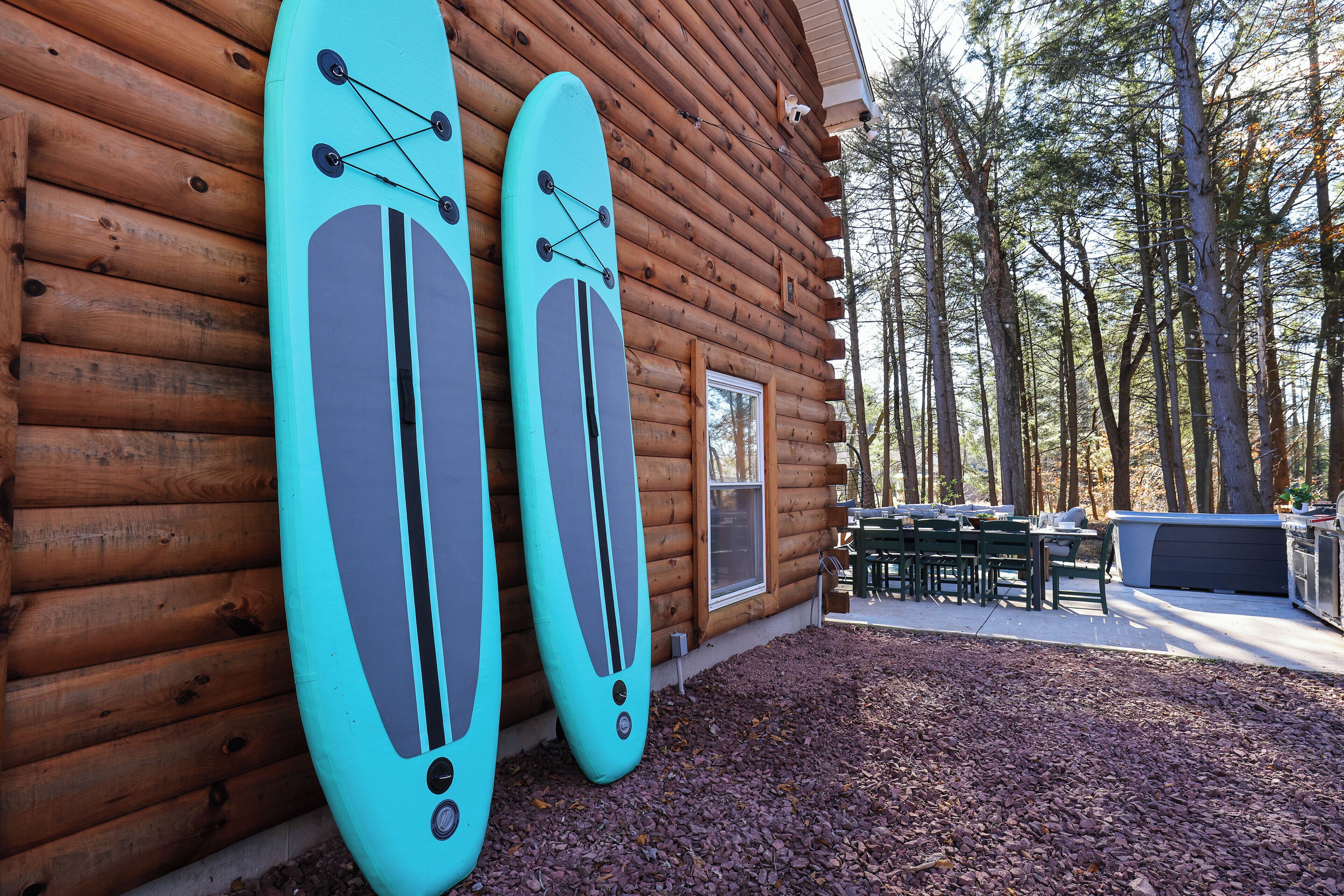 Une cabane en rondins avec deux planches de surf appuyées contre le mur, une terrasse avec des sièges d’extérieur et un paysage boisé en arrière-plan.