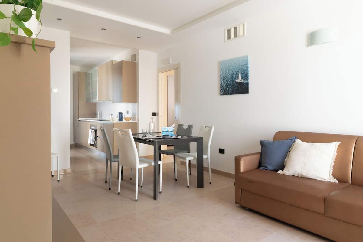 Ein modernes Wohnzimmer mit einem braunen Sofa, einem Esstisch mit Stühlen und einer Küche im Hintergrund.