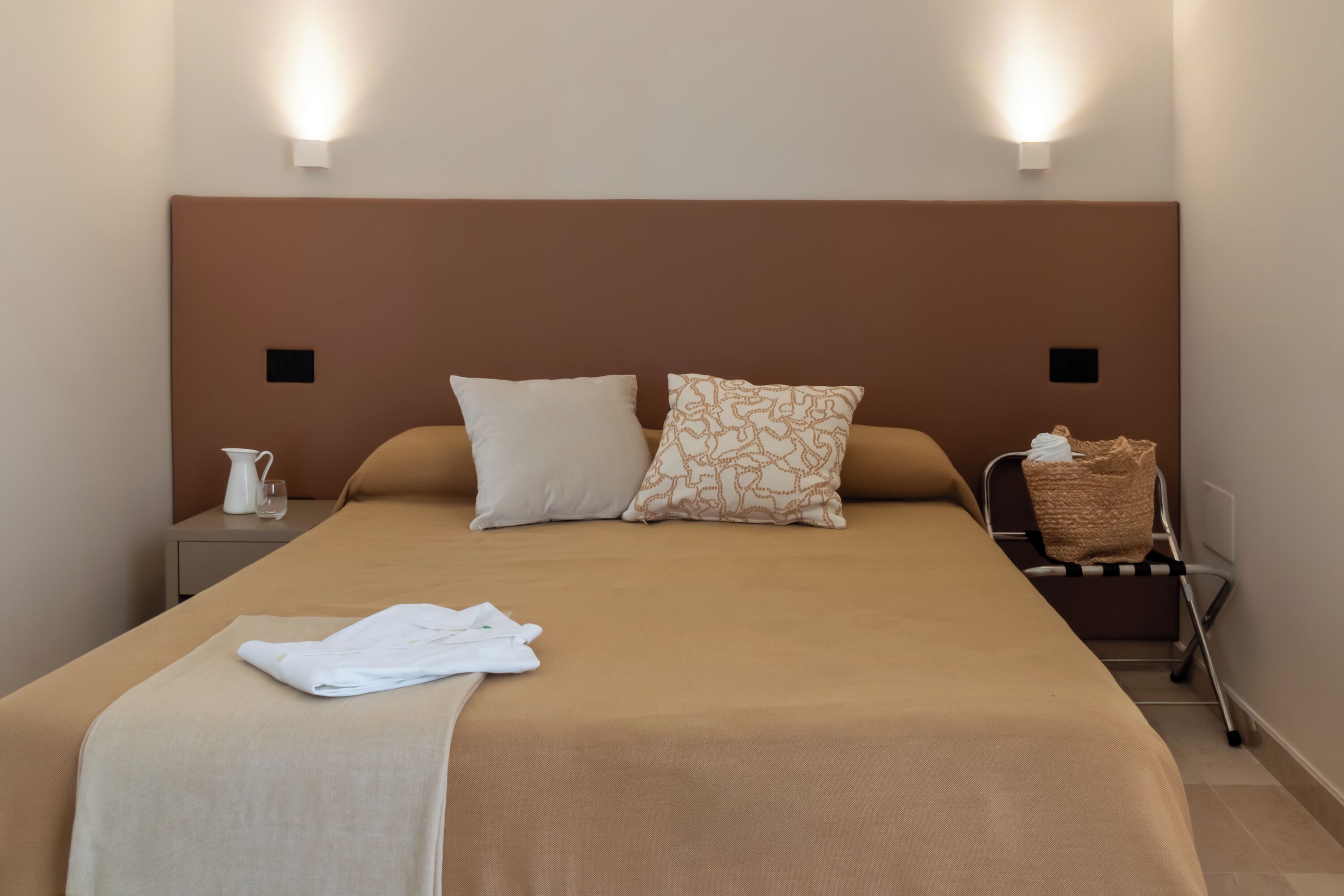 A hotel room with a large bed, two bedside lamps, a nightstand with a pitcher and glass, and a luggage trolley.