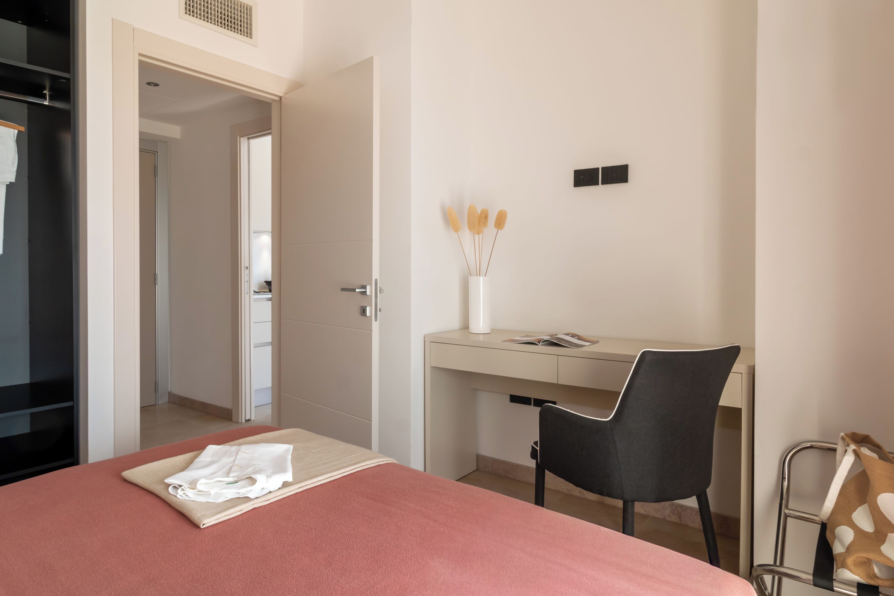A hotel room with a bed, a desk, a chair, and a small table with a vase of dried flowers.