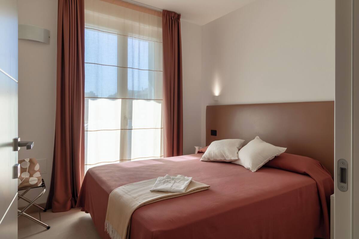 Ein Hotelzimmer mit einem großen Bett, einem Nachttisch, einem Sessel und einem Fenster mit Vorhängen.