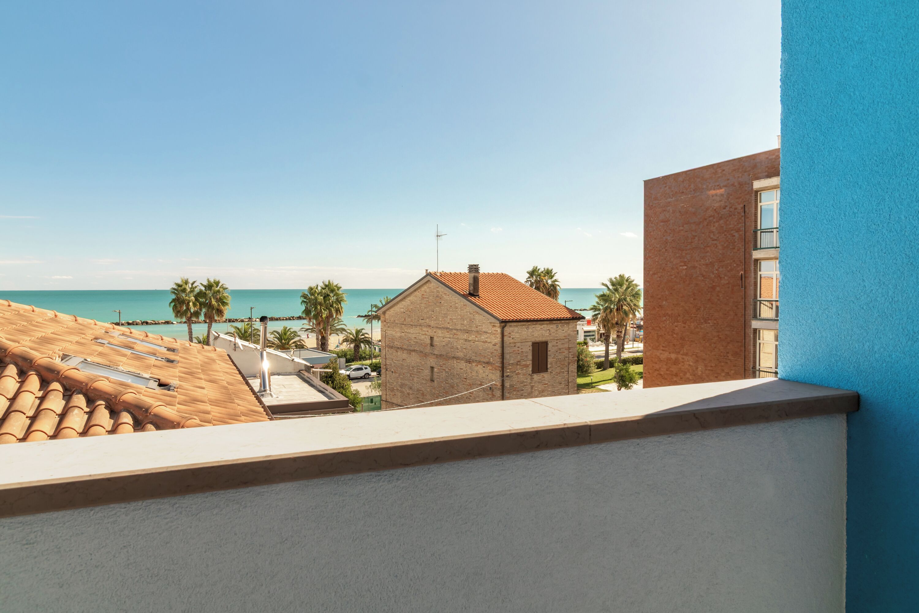 A balcony with a view of a beach, a small brick building, and a blue wall.