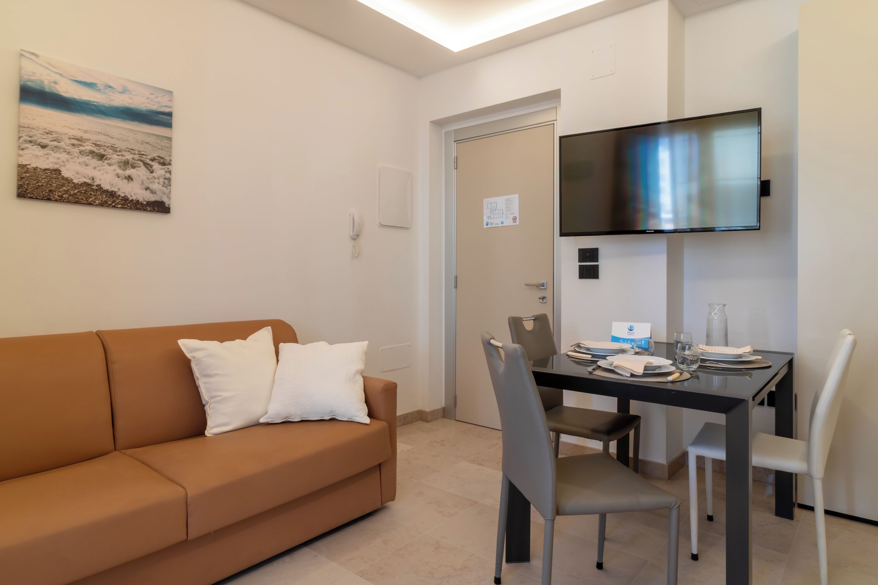 A modern living room with a brown sofa, a dining table with chairs, a flat-screen TV mounted on the wall, and a painting of a beach scene.