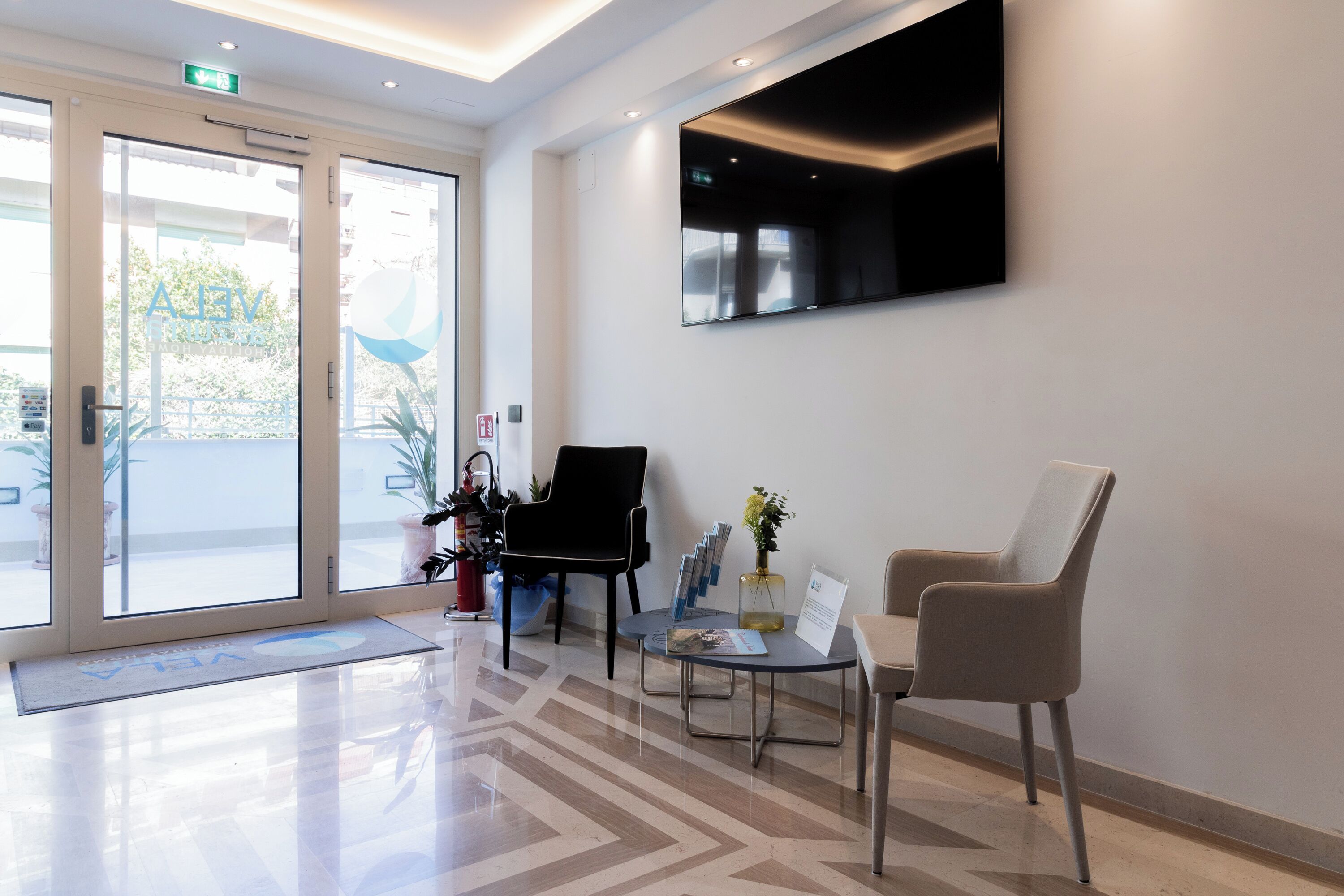 A reception area with a modern design, featuring a flat-screen TV, two chairs, a small table with a vase of flowers, and a glass door leading outside.