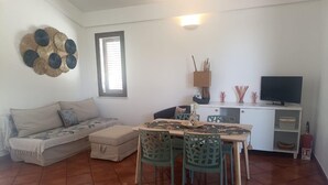 A living room with a sofa, ottoman, dining table with chairs, a television, and a cabinet. - Villa Uzzo -Scopello (Sicilia)