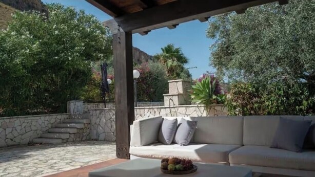 A covered outdoor seating area with a stone floor, a white sofa with cushions, and a fruit bowl on a table. - Villa Uzzo -Scopello (Sicilia)