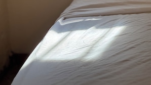Una cama con sábana blanca en una habitación con un estante de madera que sostiene una vela y un objeto pequeño.