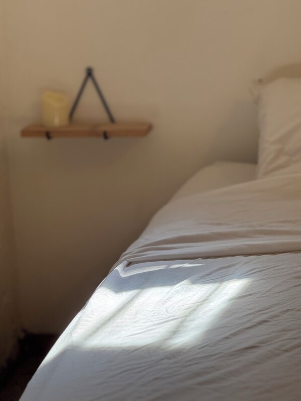A bed with a white sheet in a room with a wooden shelf holding a candle and a small item.