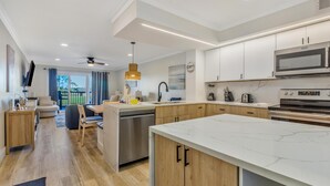 A modern kitchen with white cabinets, a large island, stainless steel appliances, and a dining area with a view of the outdoors. - Charming 1-Bedroom Harbour Point Retreat at Sandestin Golf and Beach Resort! (Miramar Beach)