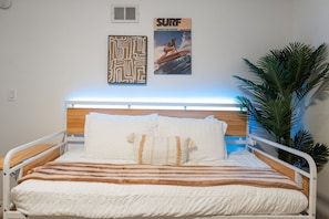 A bed with a wooden headboard, white bedding, and a striped blanket. There are two posters on the wall, one with a maze pattern and another with a surfer. - The Wave by Coastline Vacation Rentals (San Diego)