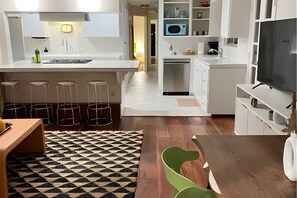 A modern kitchen with white cabinetry, a central island, bar stools, a dining table with chairs, and a television. - The Wave by Coastline Vacation Rentals (San Diego)