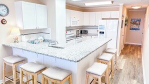 A modern kitchen with white cabinets, a granite countertop, and a central island with bar stools. - Experience Tilghman Beach and Racquet Club Unit 306! Oceanfront, Pools, Tennis, Close to Main Street! (North Myrtle Beach)