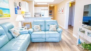 Una sala de estar con un sofá azul, una mesita con una lámpara, una cocina al fondo y un televisor sobre un mueble.