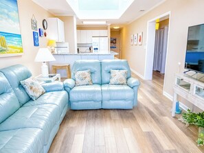 Ein Wohnzimmer mit blauem Sofa, einem kleinen Tisch mit Lampe, einer Küche im Hintergrund und einem Fernseher auf einem Schrank.