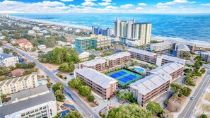 Luchtbeeld van een hotelcomplex aan de kust, met meerdere gebouwen, een tennisbaan en een strand op de achtergrond.