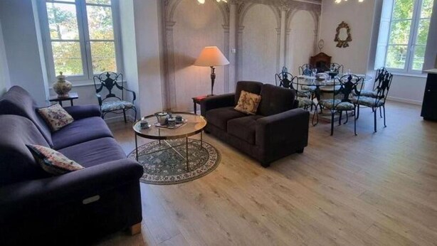 Ein geräumiges Wohnzimmer mit Sofa, Sessel und rundem Couchtisch. Es gibt einen Esstisch mit Stühlen und eine Pendelleuchte.