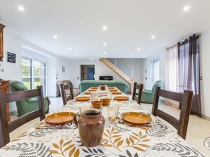 Ein Essbereich mit einem langen Tisch, gedeckt für vier Personen, umgeben von grünen Sofas. Im Hintergrund ist eine Treppe zu einer oberen Ebene zu sehen.