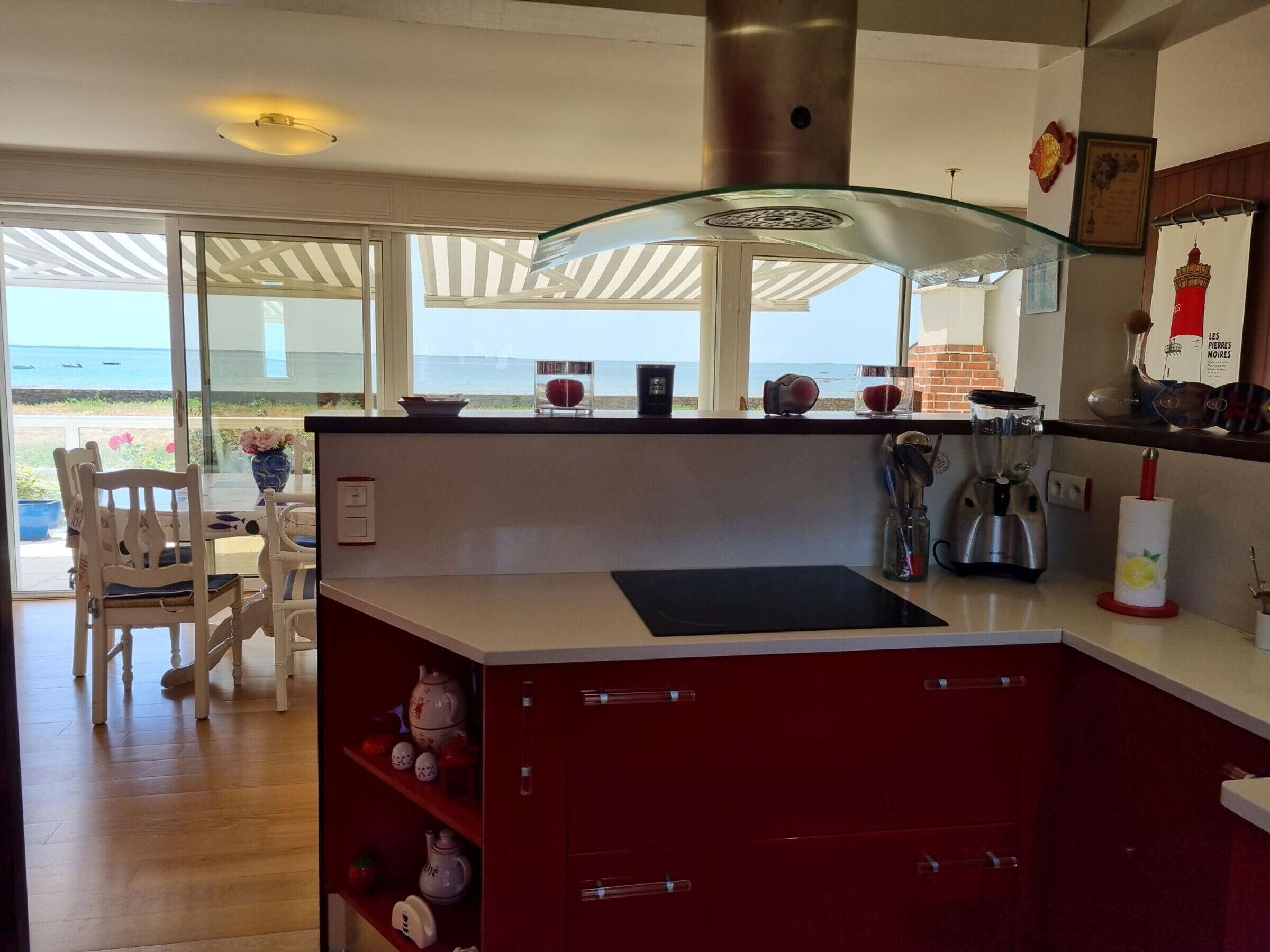 Une cuisine moderne dotée d’un grand îlot central, d’un coin repas offrant une vue sur la mer et d’un meuble rouge contenant des ustensiles de cuisine.