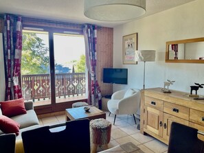 A room with a sofa, a chair, a TV, a wooden cabinet, and a balcony with a railing. - 2-bedroom apartment, ski-in/ski-out, parking, sleeps 6 (Les Carroz d'Arâches)