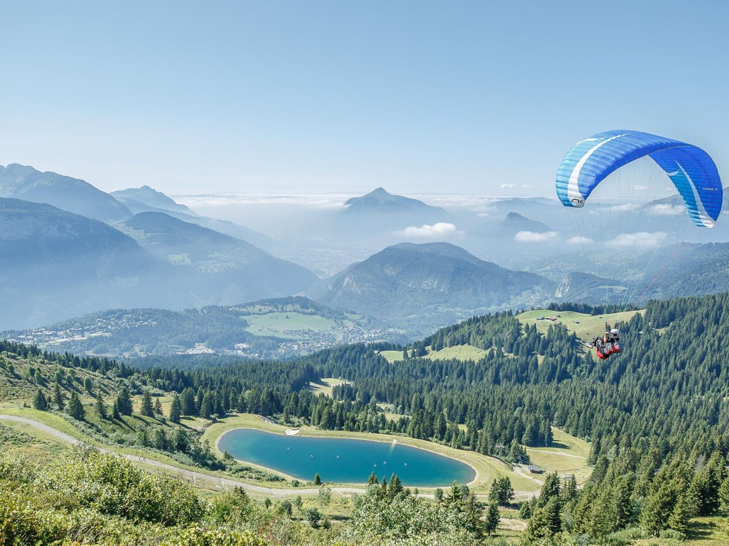 A mountainous landscape with a lake, forested areas, and a paraglider.