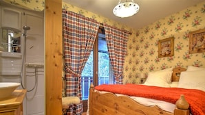 Un dormitorio con una cama de madera, una ventana con cortinas escocesas, suelo de madera y una zona de ducha con un lavabo blanco.