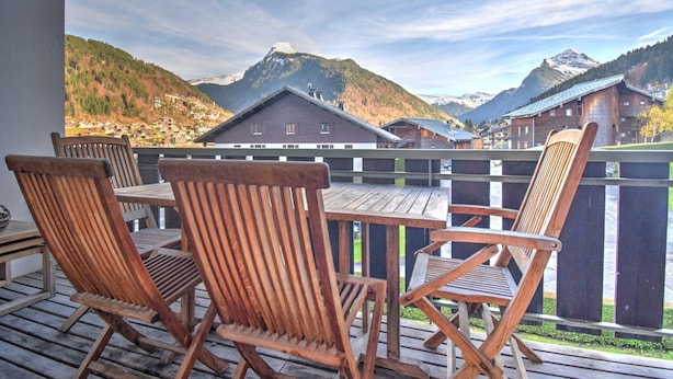 Una terraza de madera con sillas y una mesa, con vistas a un paisaje montañoso y edificios.