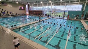 A large indoor swimming pool with multiple lanes, a scoreboard, and a view of a sports field through large windows.