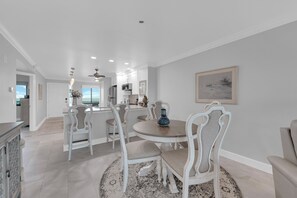 A dining area with a round table, white chairs, and a blue vase. There is a kitchen in the background with white cabinets and a refrigerator. - 2B/2B HARBOUR POINTE CONDO 816S ON THE BAY, WATCH THE BOATS GO BY, FISHING DOCK, WALK TO BEACH! (Fort Myers Beach)