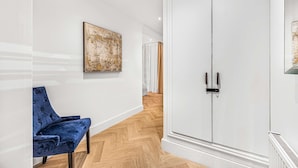 Een hal met een blauwe gestoffeerde stoel, een witte garderobe en een houten vloer met een herringbone patroon.