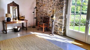A room with a stone wall, a wooden chair, a desk with a lamp, and a large window with a view of greenery. - Charming Basque house with swimming pool (Souraïde)
