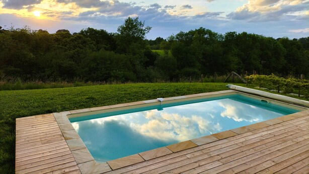 A rectangular swimming pool with a wooden deck, surrounded by lush greenery and trees, under a partly cloudy sky. - Charming Basque house with swimming pool (Souraïde)