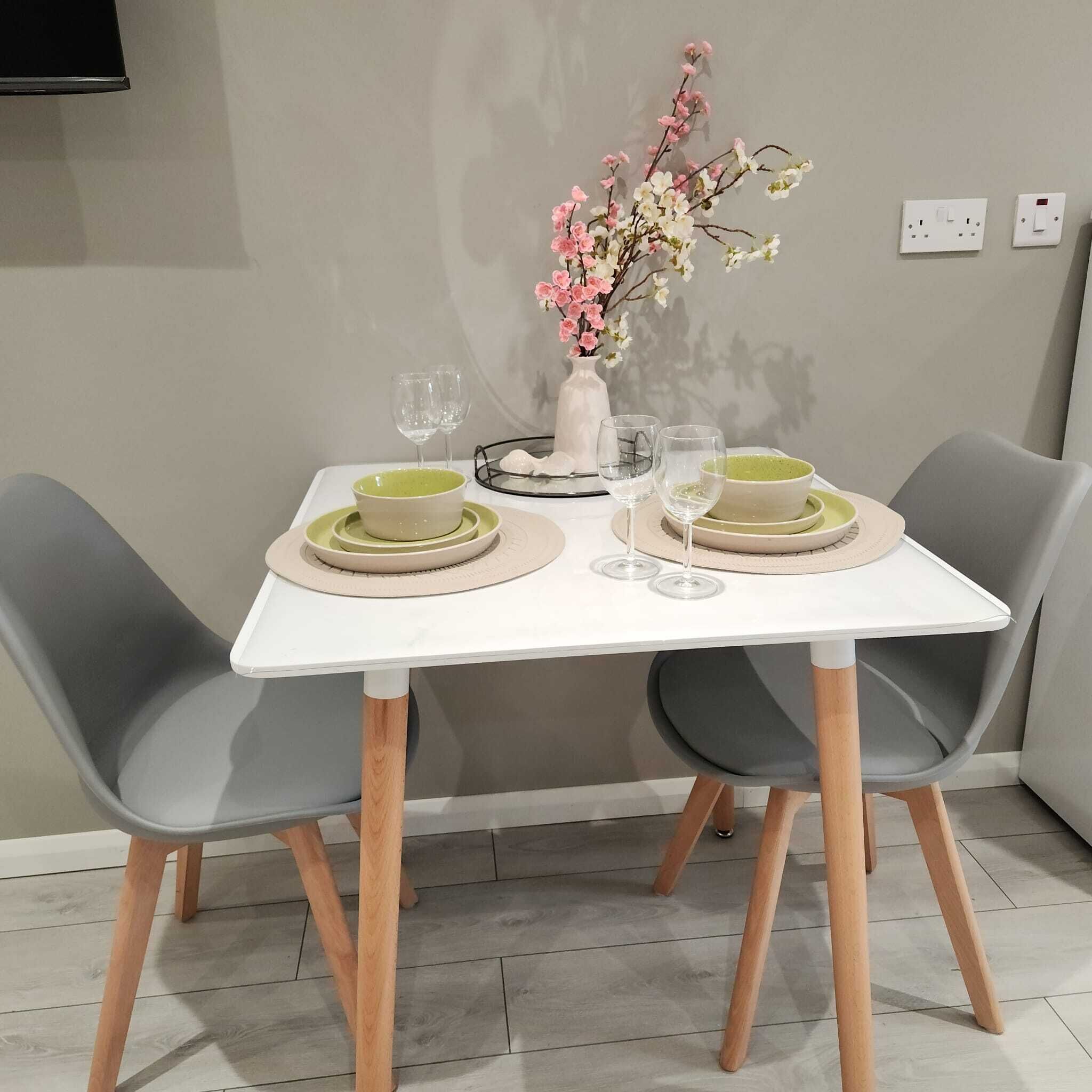 A dining table with two chairs, a vase with flowers, and two sets of dinnerware.