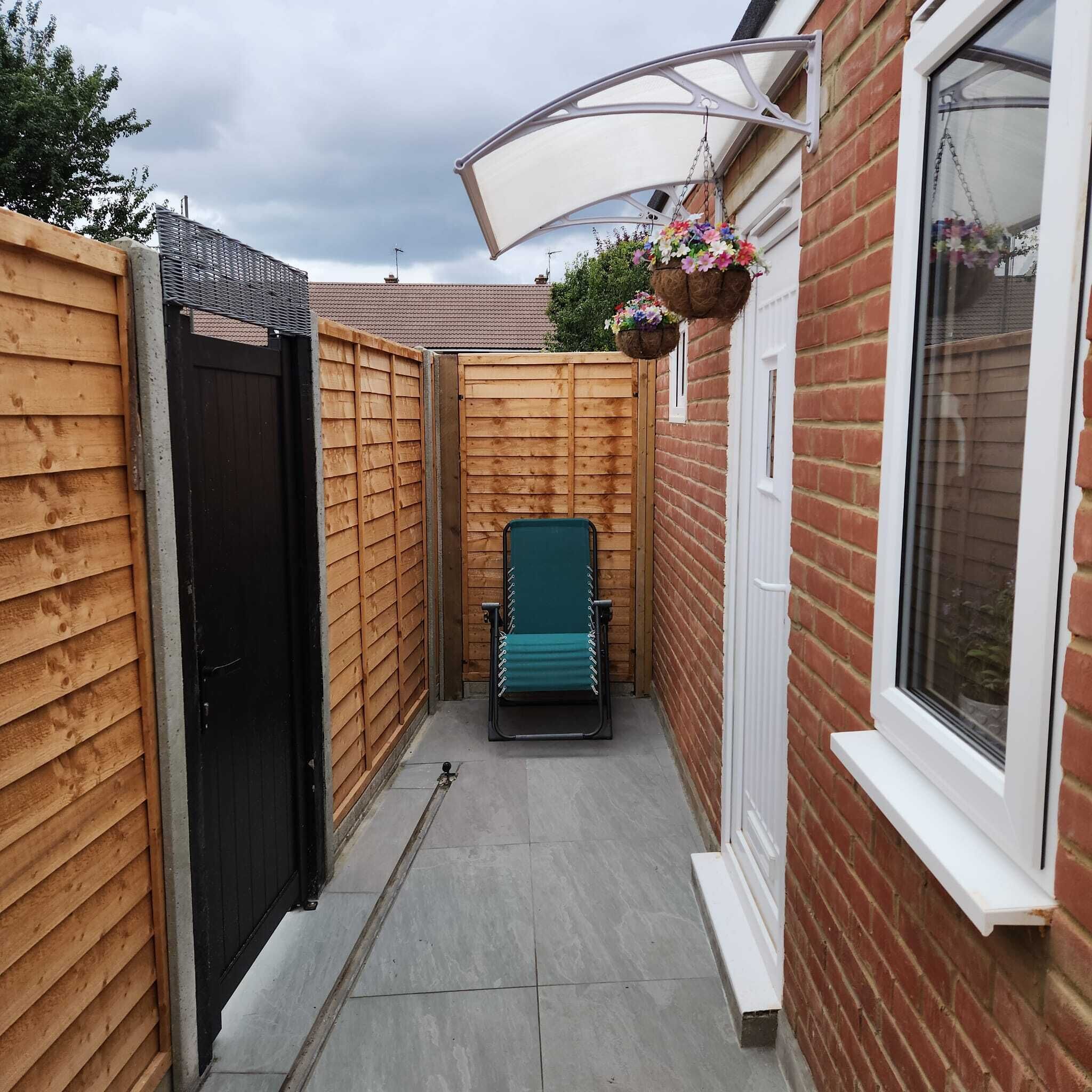 Un étroit couloir extérieur avec une chaise verte, des murs en bois, une porte noire et un pot de fleurs suspendu.