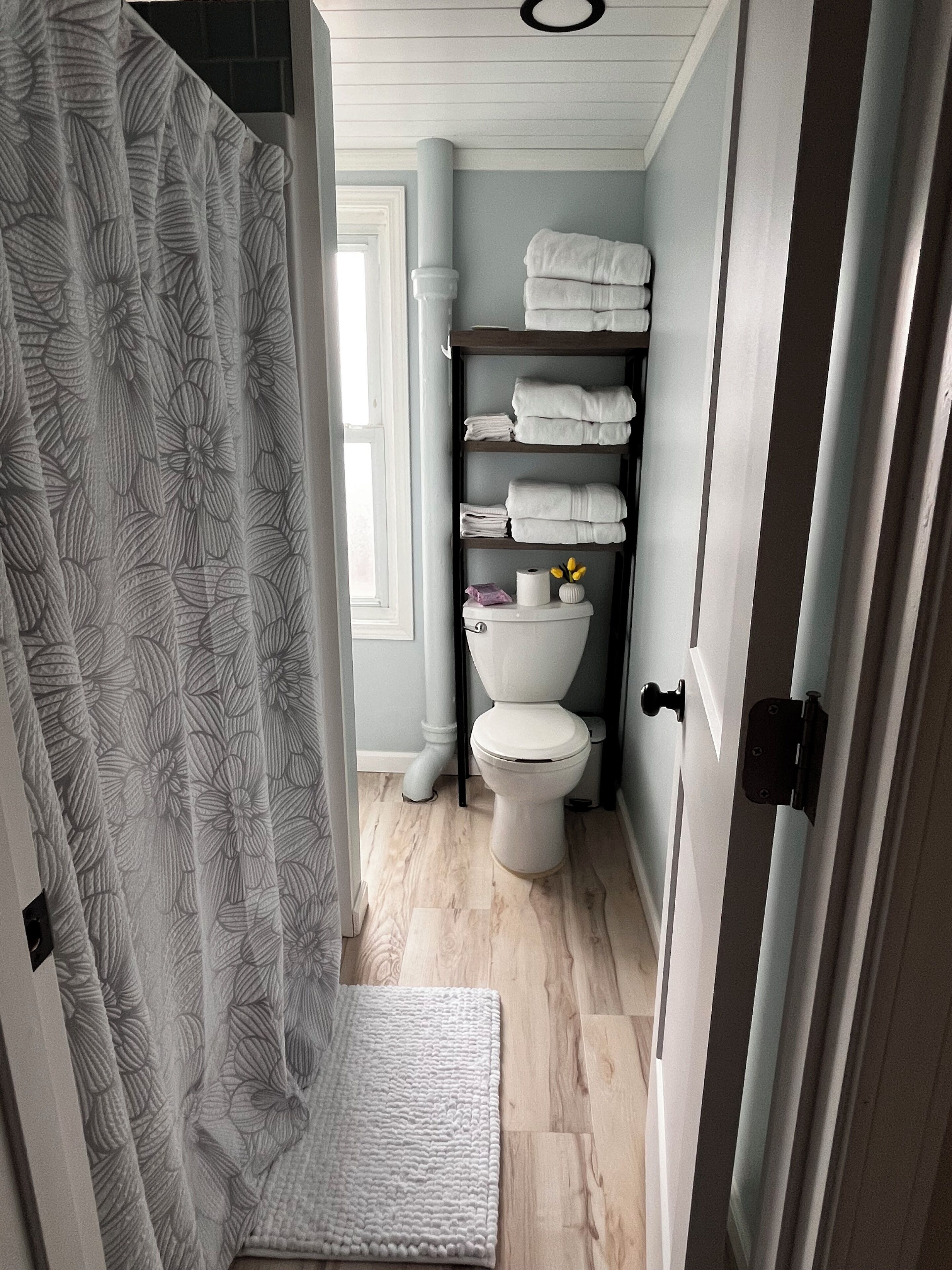 A bathroom with a shower curtain, a toilet, a shelf with towels, and a small window.