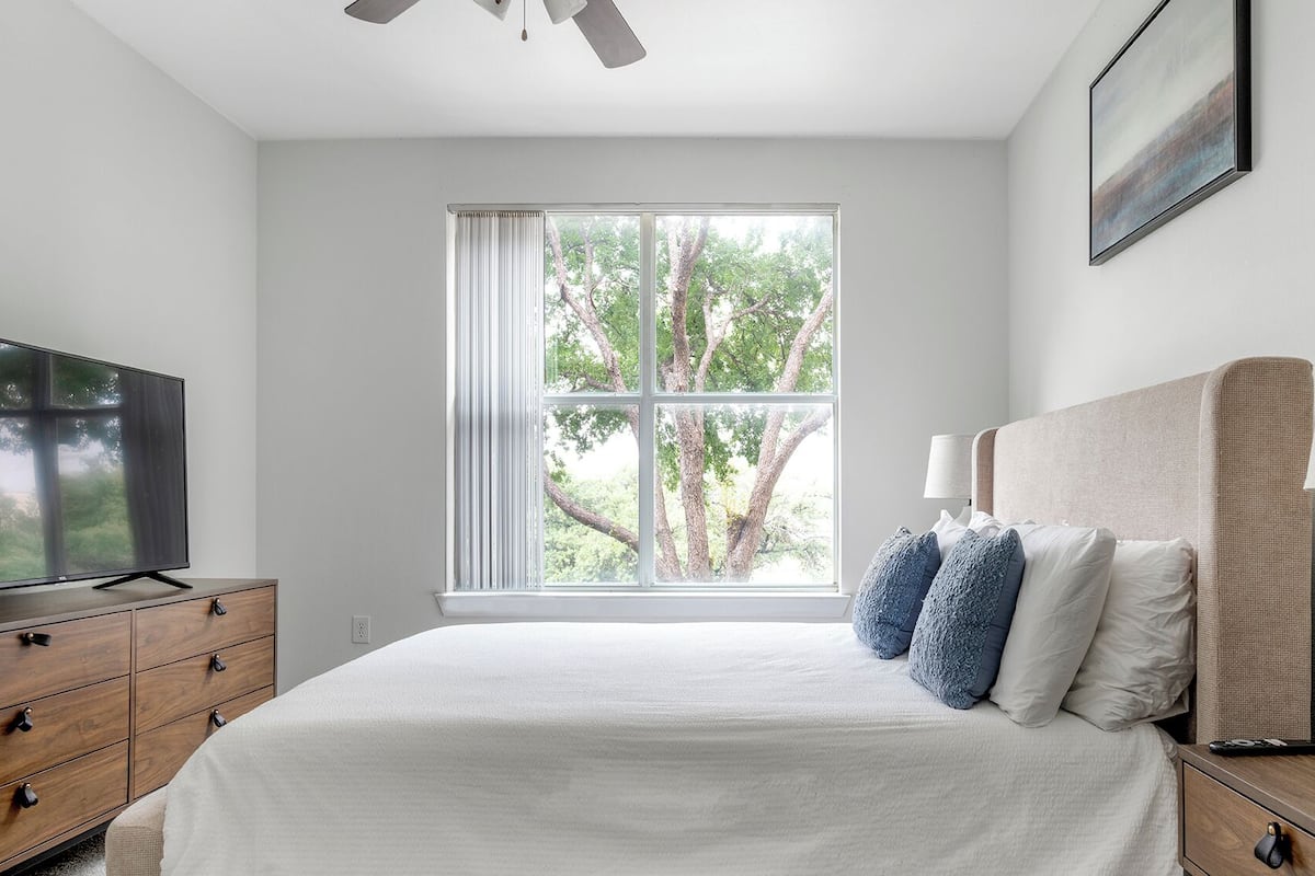 A bedroom with a large window, a bed with pillows, a TV on a wooden dresser, and a lamp on a nightstand.