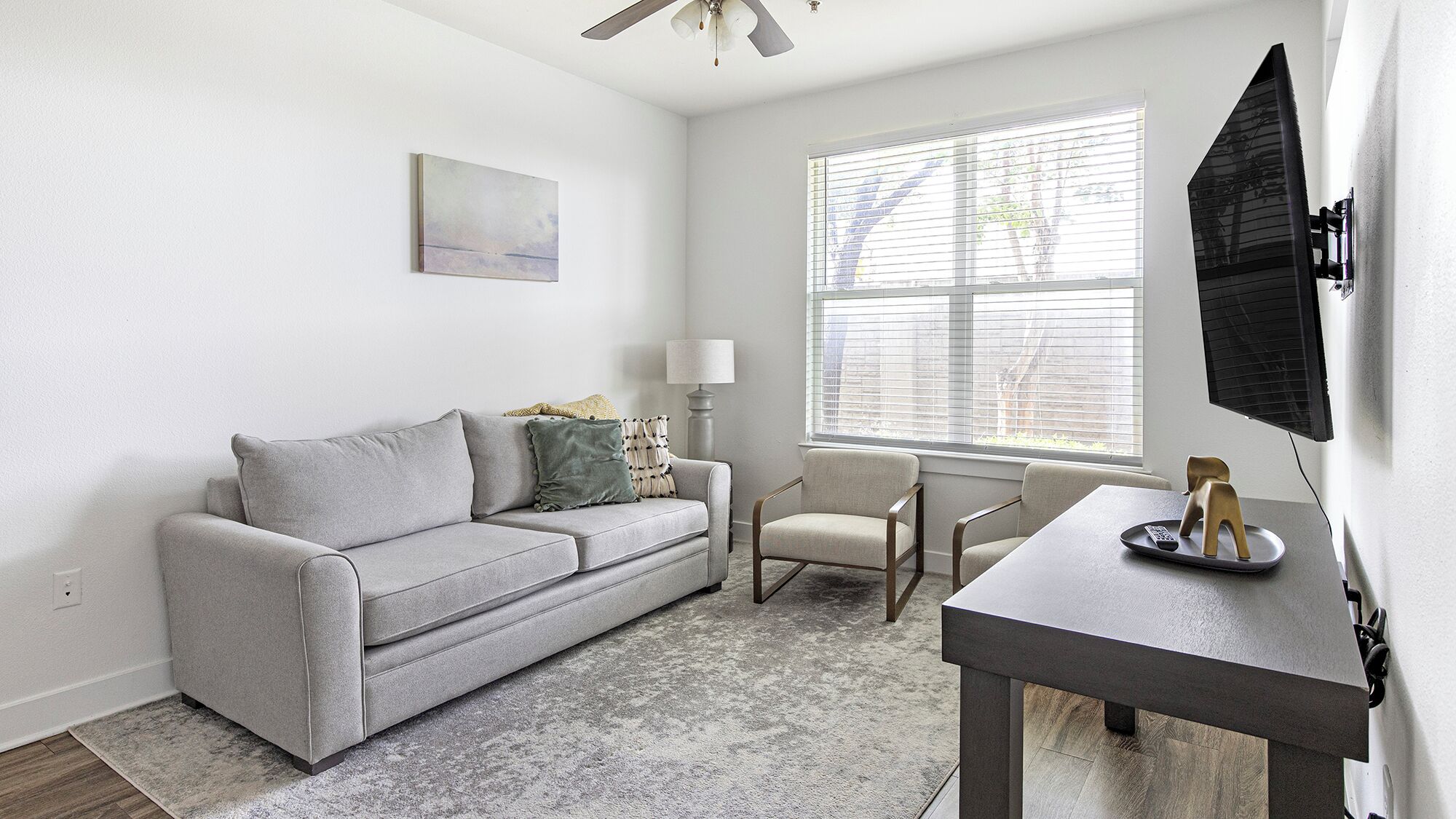 A living room with a grey sofa, a small table, a chair, a lamp, and a TV mounted on the wall.
