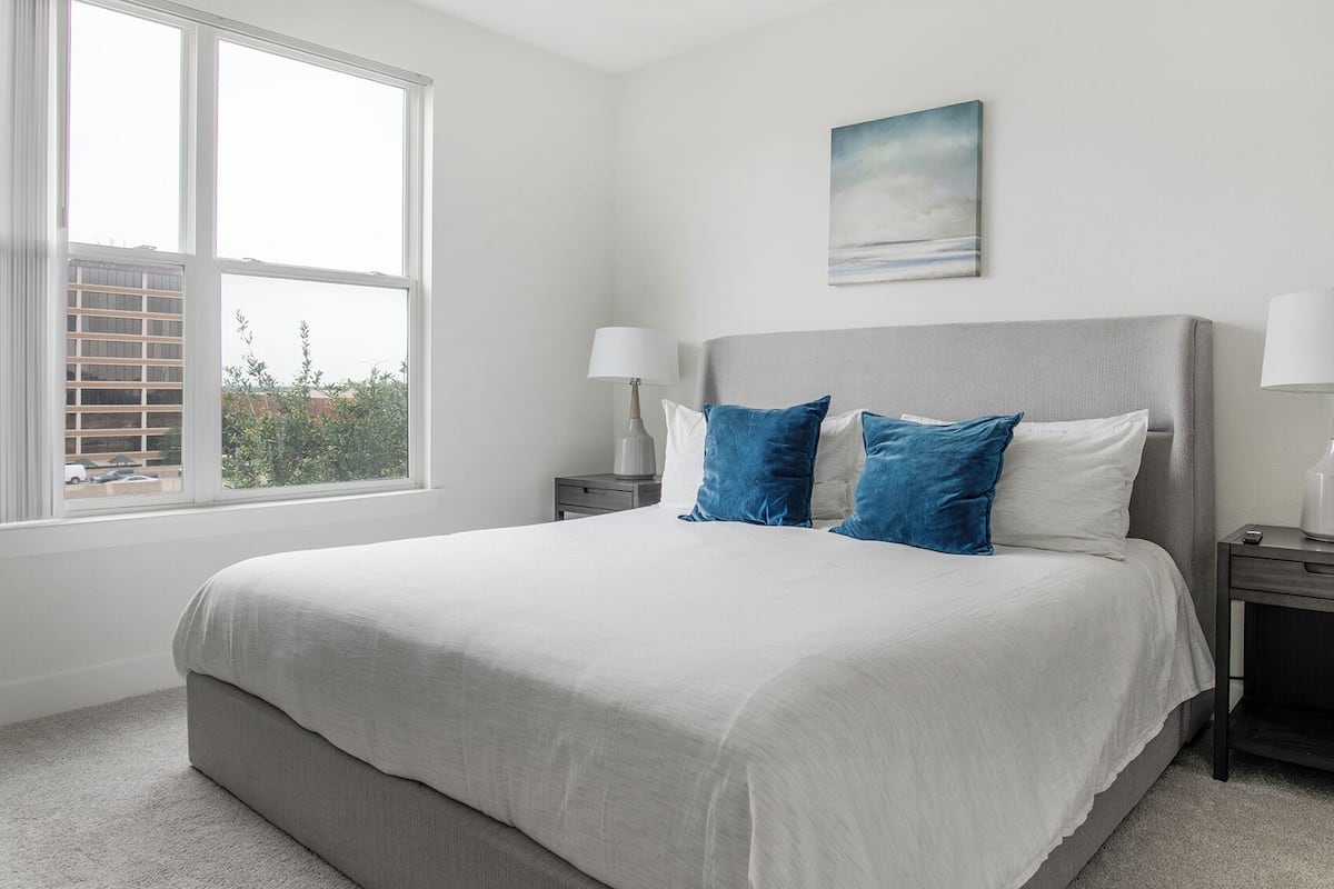 A bedroom with a large bed, two bedside tables, a window with a view of buildings, and a painting on the wall.
