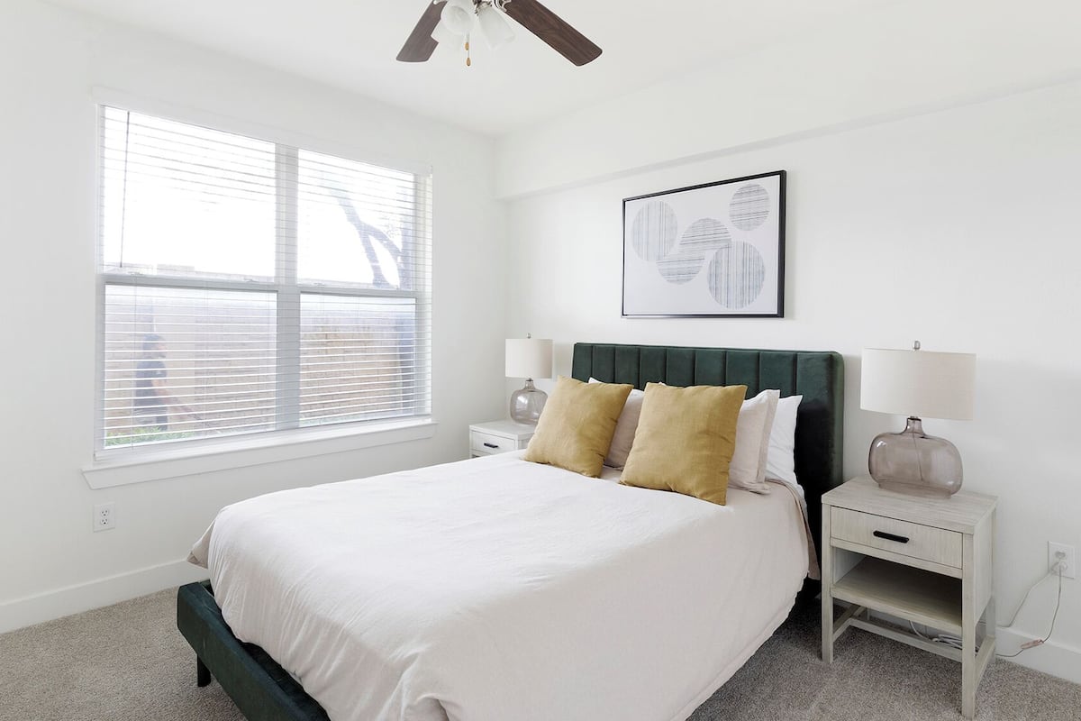 A bedroom with a bed, two bedside tables, a ceiling fan, a window with blinds, and a framed picture on the wall.