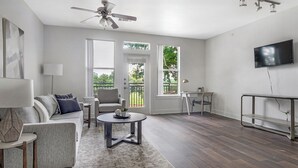 A modern living room with a ceiling fan, a flat-screen TV mounted on the wall, a sofa with cushions, a coffee table, and a desk with a chair. - Landing Apartments - North Dallas (Dallas)