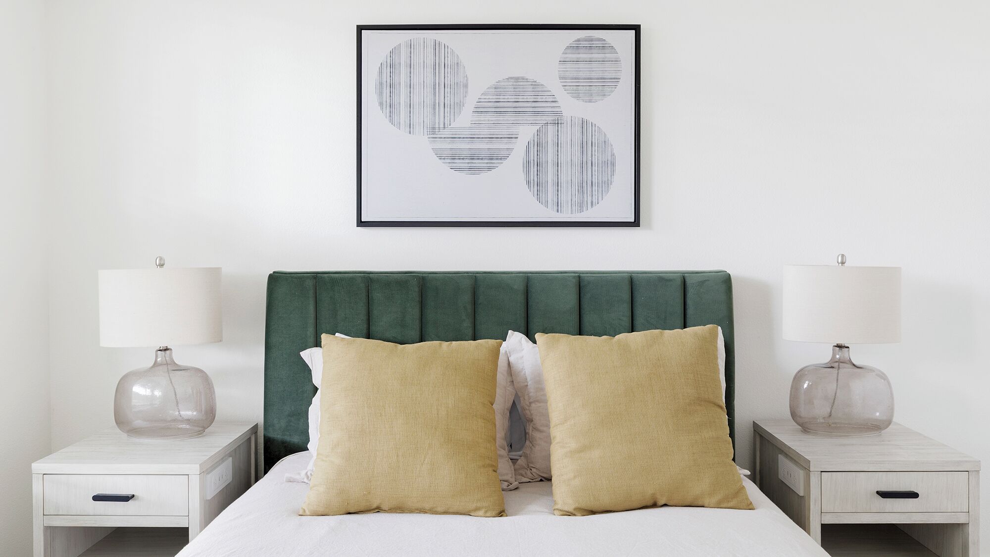 Una camera da letto ordinata con una testiera imbottita verde, due comodini con lampade e un quadro astratto appeso sopra il letto.