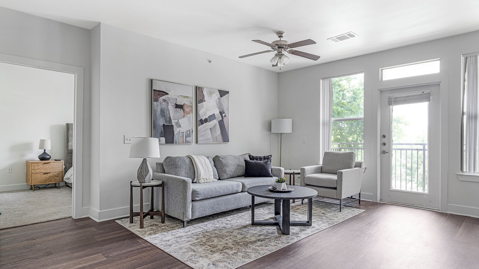 A modern living room with a grey sofa, a coffee table, a side table with a lamp, and a ceiling fan. There are two windows with curtains, a door, and a small side table with a lamp.