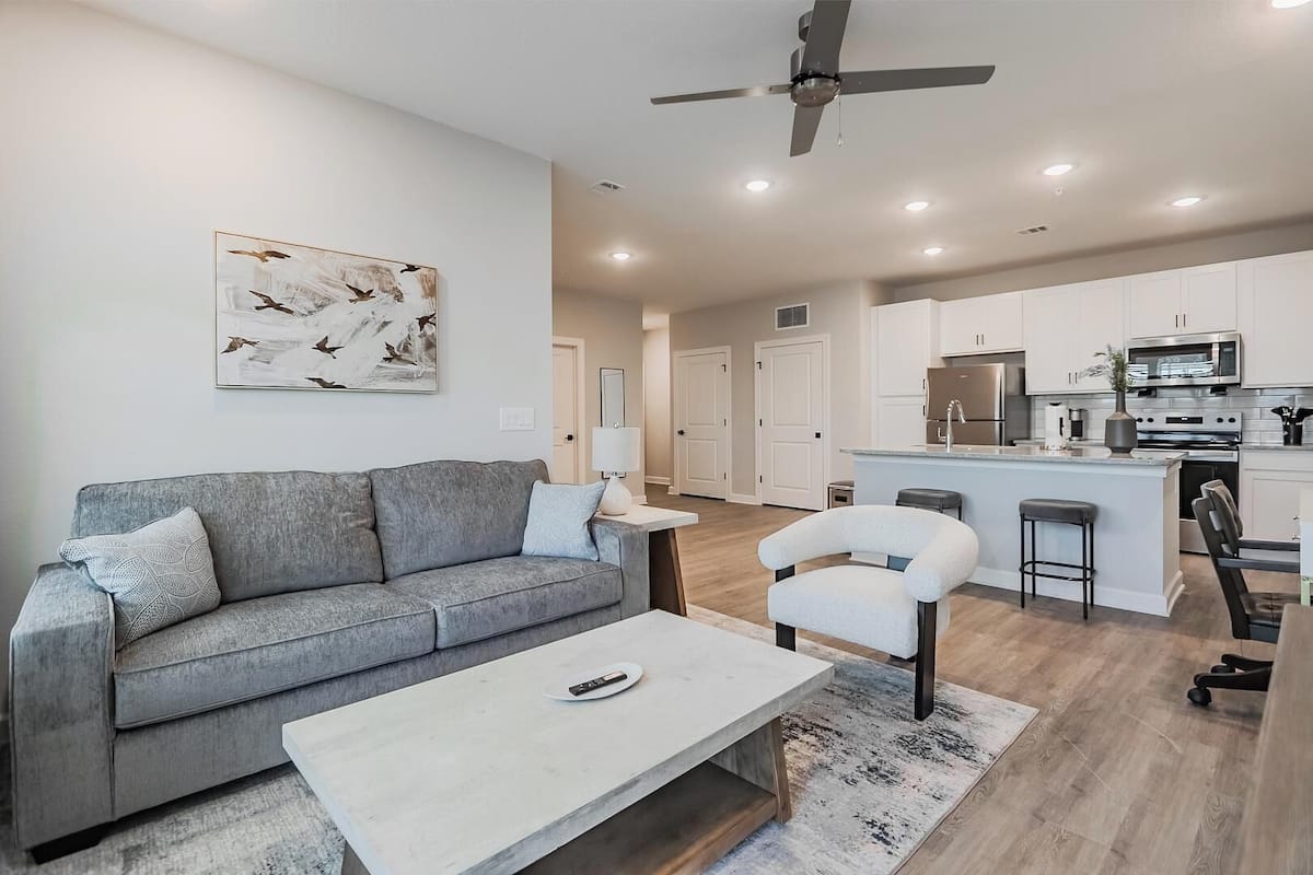 A modern living room with a grey sofa, a white armchair, and a coffee table. A ceiling fan is visible, and the room has a neutral color palette.